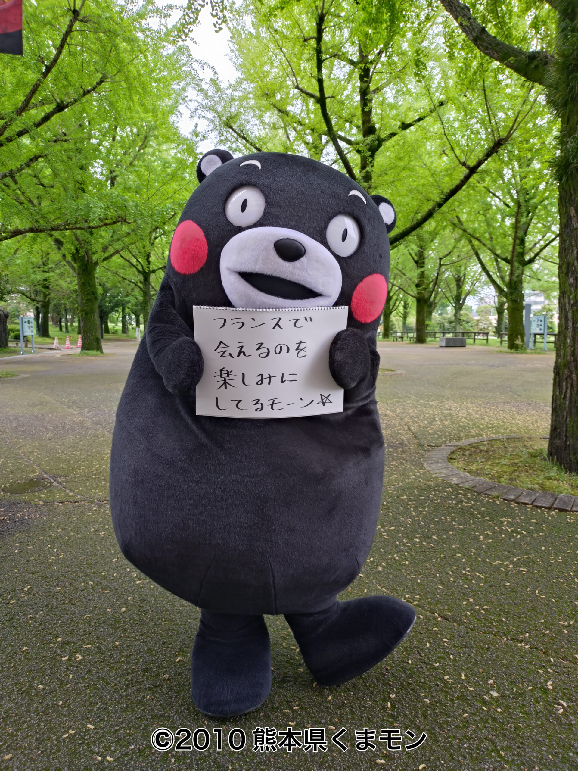 片足を上げたくまモンが公園で「フランスで会えるのを楽しみにしてるモーン☆」のメッセージを掲げている様子。
